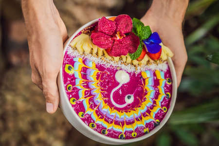 Young man having a mediterranean breakfast, eats Healthy tropical breakfast, smoothie bowl with tropical fruits, decorated with a pattern of colorful yogurt with turmeric and spirulina. It is also decorated with fruits, flowers, chia seeds, coconut, granola, pineapple, mint and strawberries.の写真素材