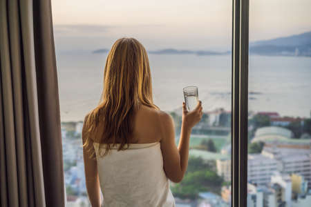 Woman drinks water in the morning on a background of a window with a sea view.