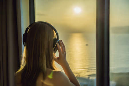 Woman in the morning listens to wireless headphones on a background of a window with a sea view.の写真素材