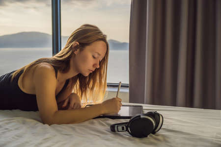 Young woman draws on a tablet against the background of a window with a sea view. Designer,...