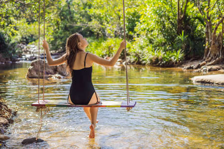 Beautiful happy girl, young pretty cheerful woman traveler sitting, swinging on a swing tied to a tree above the water, smiling in sunny day. Summer vacation, travel concept.の写真素材
