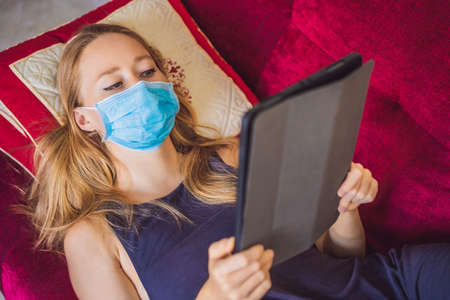 Woman in medical mask reads a book on the couch. Home quarantine. Stay at home. Coronovirus