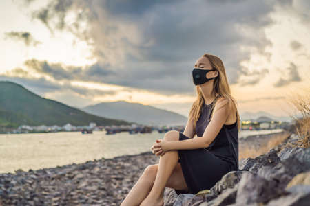 Self-isolation in nature. Young woman in Fashionable black medical mask with filter sitting by the river. Coronavirus 2019-ncov epidemic concept. Woman in a black medical mask. Portrait of a woman with expressive eyes during a virus or disease epidemic of Coronovirusの写真素材