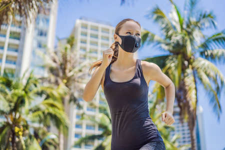 Woman runner wearing medical mask. Running in the city against the backdrop of the city. Coronavirus pandemic Covid-19. Sport, Active life in quarantine surgical sterilizing face mask protection. Outdoor run on athletics track in Corona Outbreakの写真素材