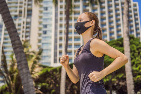 Woman runner wearing medical mask. Running in the city against the backdrop of the city. Coronavirus pandemic Covid-19. Sport, Active life in quarantine surgical sterilizing face mask protection. Outdoor run on athletics track in Corona Outbreakの写真素材