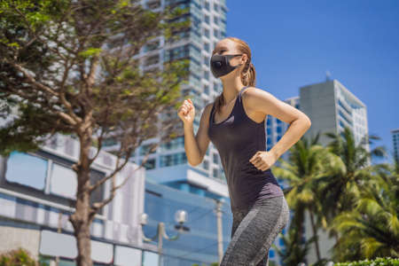Woman runner wearing medical mask. Running in the city against the backdrop of the city. Coronavirus pandemic Covid-19. Sport, Active life in quarantine surgical sterilizing face mask protection. Outdoor run on athletics track in Corona Outbreakの写真素材