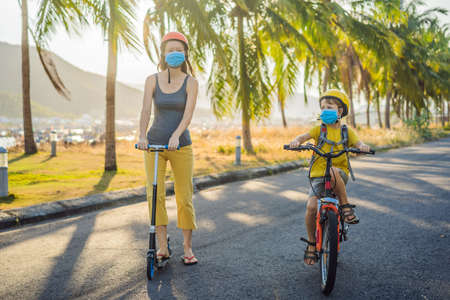 Active school kid boy and his mom in medical mask and safety helmet riding a bike with backpack on...