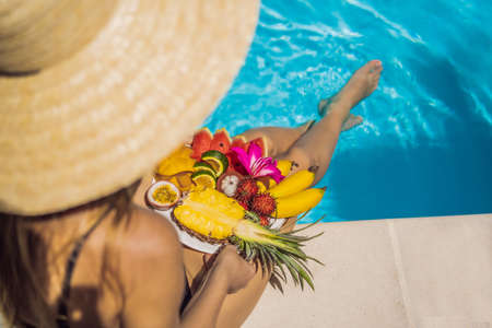 Young woman relaxing and eating fruit plate by the hotel pool. Exotic summer diet. Photo of legs with healthy food by the poolside, top view from above. Tropical beach lifestyleの写真素材