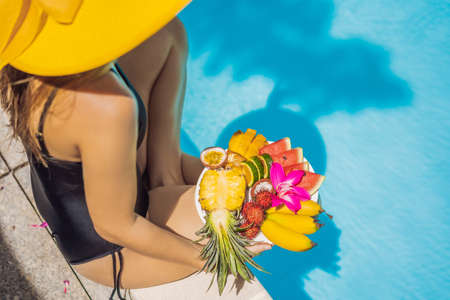 Young woman relaxing and eating fruit plate by the hotel pool. Exotic summer diet. Photo of legs with healthy food by the poolside, top view from above. Tropical beach lifestyleの写真素材