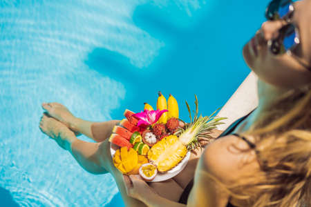 Young woman relaxing and eating fruit plate by the hotel pool. Exotic summer diet. Photo of legs with healthy food by the poolside, top view from above. Tropical beach lifestyleの写真素材