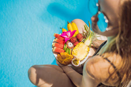 Young woman relaxing and eating fruit plate by the hotel pool. Exotic summer diet. Photo of legs with healthy food by the poolside, top view from above. Tropical beach lifestyleの写真素材