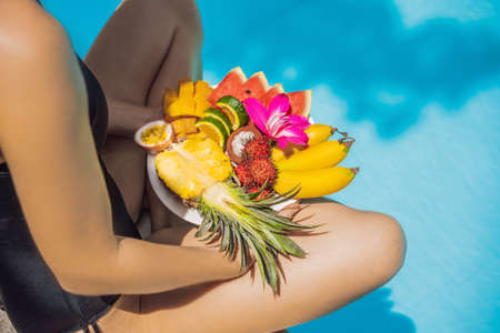 Young woman relaxing and eating fruit plate by the hotel pool. Exotic summer diet. Photo of legs with healthy food by the poolside, top view from above. Tropical beach lifestyleの写真素材