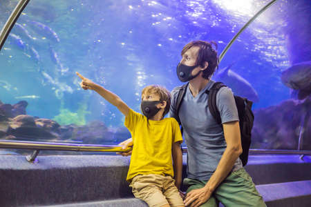 Dad and son in medical masks look at the fish in the aquarium in oceanarium. People walk in medical masks after the end of the coronovirusの写真素材