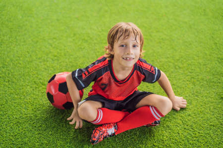 Little cute kid boy in red football uniform playing soccer, football on field, outdoors. Active child making sports with kids or father, Smiling happy boy having fun in summerの写真素材