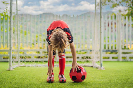 Little cute kid boy in red football uniform playing soccer, football on field, outdoors. Active child making sports with kids or father, Smiling happy boy having fun in summerの写真素材