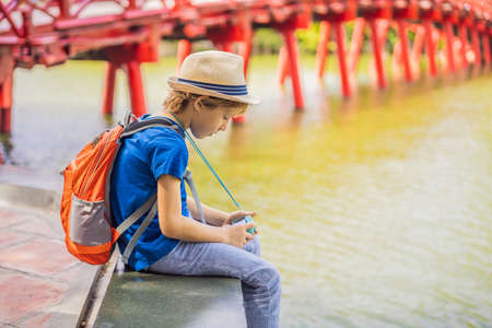 Caucasian boy tourist on background of Red Bridge in public park garden with trees and reflection in the middle of Hoan Kiem Lake in Downtown Hanoi. Traveling with children conceptの写真素材