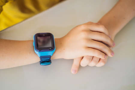 Little boy sitting at the table and looking smart watch. Smart watch for baby safety. The child makes school lessons, listening to music, calling friendsの写真素材