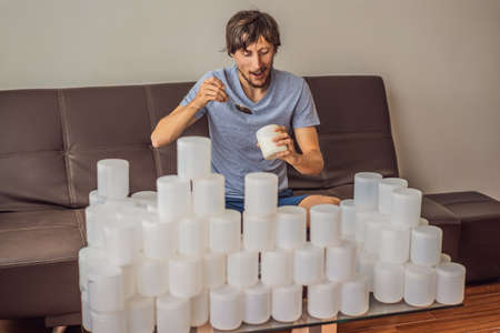 The man eats yogurt and is surrounded by many jars of yoghurt that he uses in a year. A huge amount of plastic food jars. Excessive consumption of plastic. Reduce, reuse, recycleの写真素材