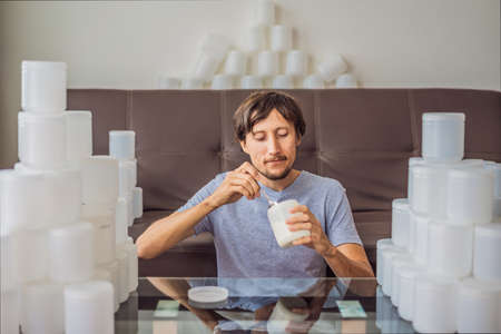 The man eats yogurt and is surrounded by many jars of yoghurt that he uses in a year. A huge amount of plastic food jars. Excessive consumption of plastic. Reduce, reuse, recycleの写真素材