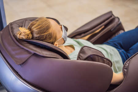 Beautiful young woman wearing a medical mask due to the coronavirus COVID 19 relaxing on the massage chair in airport or in the mallの写真素材