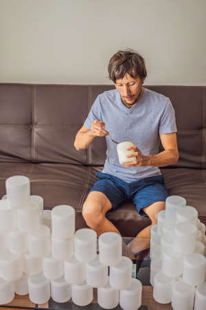 The man eats yogurt and is surrounded by many jars of yoghurt that he uses in a year. A huge amount of plastic food jars. Excessive consumption of plastic. Reduce, reuse, recycleの写真素材
