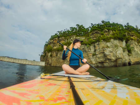 Young women Having Fun Stand Up Paddling in the sea. SUP. Red hair girl Training on Paddle Board near the rocksの写真素材