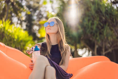 Summer lifestyle portrait of pretty girl sitting on the orange inflatable sofa and drinking water on the beach of tropical island. Relaxing and enjoying life on air bedの写真素材