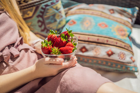 Portrait of gorgeous woman wearing beautiful dress sitting in a patio and eating strawberryの写真素材