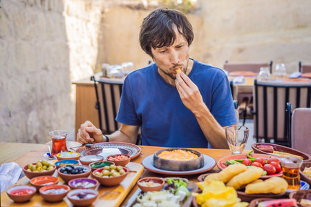 Man eating turkish breakfast. Turkish breakfast table. Pastries, vegetables, greens, olives, cheeses, fried eggs, spices, jams, honey, tea in copper pot and tulip glasses, wide compositionの写真素材