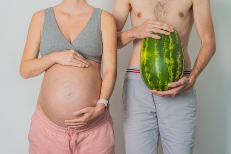 A humorous image: a pregnant woman and her husband playfully use a watermelon in place of a belly, comically highlighting the challenges of navigating with a pregnant bumpの写真素材