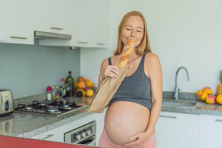 Pregnant woman eating bread in the kitchen. Exploring the impact of gluten during pregnancy: understanding the potential benefits and risks for maternal health and fetal developmentの写真素材