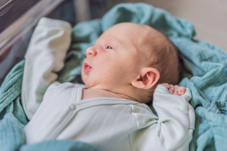 A newborn rests peacefully in his transparent bassinet in the hospital. The clear bassinet provides...