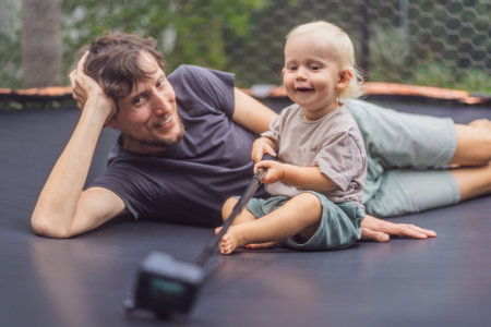 Dad and toddler sitting on an outdoor trampolinewith an action camera. Family bonding, playful moments, and outdoor fun conceptの写真素材