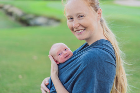 Mom walks with her newborn baby in a sling. This moment highlights the close bond between mother and child, promoting comfort, security, and the benefits of babywearing in everyday lifeの写真素材