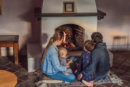 Family bonding by the fireplace during cozy winter evening. Warm and peaceful family moment indoors. Togetherness, love, and relaxation concept. A warm and inviting scene featuring a lit fireplace with glowing firewood. A colorful traditional rug and pillow lie on the floor, creating a cozy atmosphere. Home, relaxation, and rustic comfort conceptの写真素材