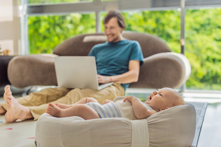 A 3-month-old baby lies safely in a cozy infant cocoon, secured with a special safety belt, while their father works on a laptop in the background. Work-life balance, parenting, and modern family conceptの写真素材
