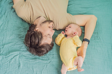 A father and his newborn baby lie on a soft muslin swaddle, sharing a tender moment of closeness and...