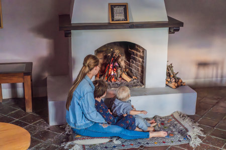 Family bonding by the fireplace during cozy winter evening. Warm and peaceful family moment indoors. Togetherness, love, and relaxation concept. A warm and inviting scene featuring a lit fireplace with glowing firewood and candles. A colorful traditional rug and pillow lie on the floor, creating a cozy atmosphere. Home, relaxation, and rustic comfort conceptの写真素材
