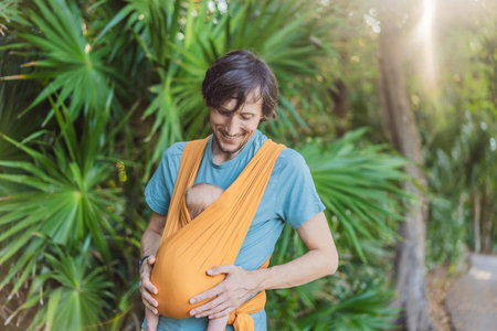 Father holding his little baby in a yellow sling in the park. Warm and loving family moment....