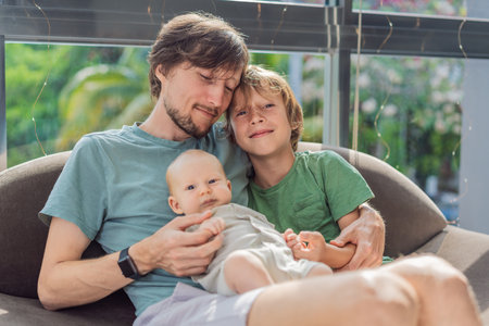Father, son, and newborn baby sitting on the couch, joyfully celebrating the newest addition to...
