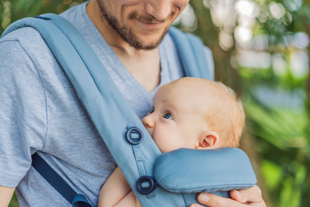 Young father walks in nature with his baby son nestled close in a parent-facing ergonomic baby...