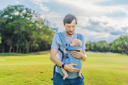 Young father walks in nature with his baby son nestled close in a parent-facing ergonomic baby...