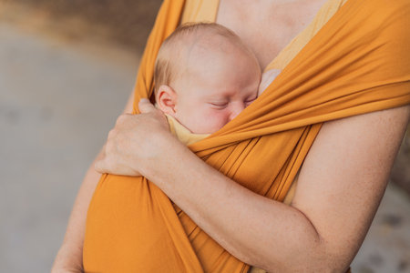 Mother holding her little baby in a yellow sling in the park. Warm and loving family moment. Babywearing, parenting, and mother-child bonding conceptの写真素材