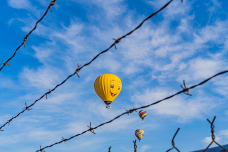 Hot air balloon with a painted smile floats in a clear blue sky, viewed through barbed wire, creating a playful yet thought-provoking travel and freedom conceptの写真素材