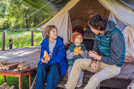 Father with his toddler and teenage son tourists in warm vests and hats eating buns while sitting on glamping steps, enjoying family travel, outdoor adventure, and bondingの写真素材