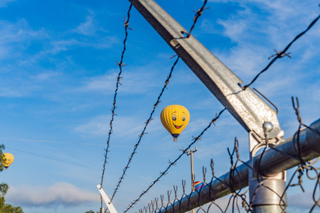 Hot air balloon with a painted smile floats in a clear blue sky, viewed through barbed wire, creating a playful yet thought-provoking travel and freedom conceptの写真素材