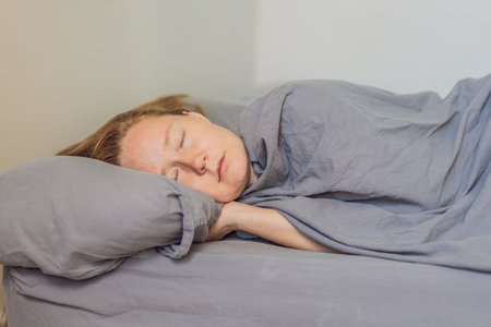Woman peacefully sleeping in bed with soft gray bedding, enjoying a calm and comfortable morning atmosphere. Rest, relaxation and wellbeing concept, representing healthy sleep, serenity and cozy home lifestyleの写真素材