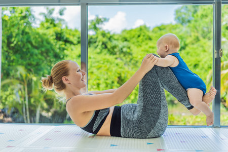 Mother doing fitness exercises while holding her baby at home, combining workout and childcare. Real...