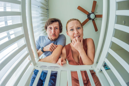 Mother and father leaning over baby crib and looking at their newborn from infant point of view. Loving parents, family bonding, childcare and emotional connection concept at home