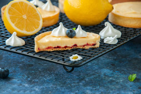 Whole and slice of individual lemon curd tarts with blueberry jam meringue and basil leaves on a blue rusty background. Flat lay and copy space.の写真素材